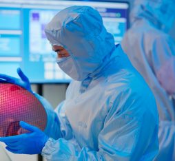 A technician in sterile coverall and gloves holds a wafer that reflects many different colors and checks it at a semiconductor manufacturing plant / Un technicien observe un wafer dans un centre de fabrication de semi-conducteurs