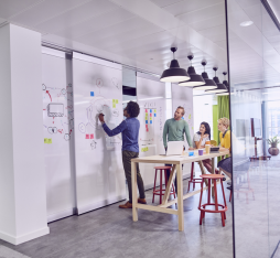 Dans un bureau moderne et lumineux, un groupe de quatre personnes est engagé dans une séance de brainstorming. Un homme écrit sur un tableau blanc, tandis que trois autres personnes, assises autour d'une table en bois, discutent et regardent un ordinateur portable. Des notes autocollantes colorées sont visibles sur le tableau. L'espace est décoré avec des rideaux colorés et des lampes suspendues, créant une atmosphère dynamique et collaborative.