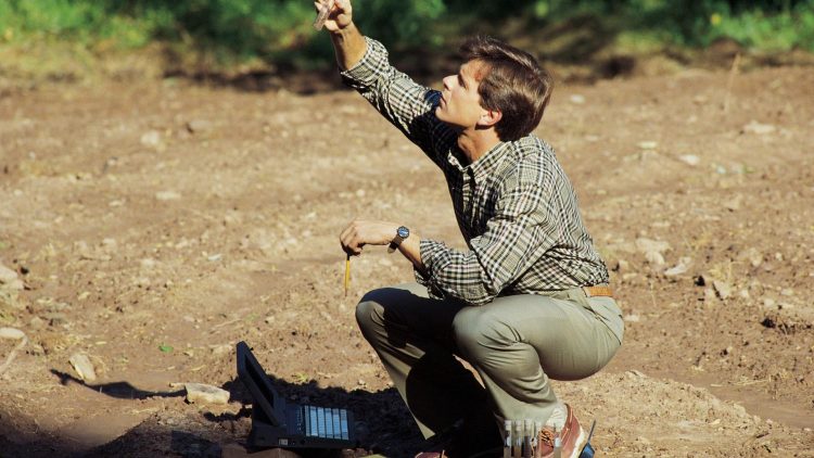 Un homme est accroupi sur un sol nu, tenant un objet en l'air avec une main et un crayon dans l'autre. À côté de lui, un ordinateur portable est ouvert, et il semble concentré sur son travail de recherche en extérieur.