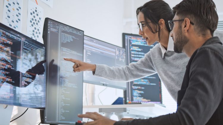 Two people collaborate in front of computer screens, one pointing something out to the other. The screens display computer code in a modern office environment.