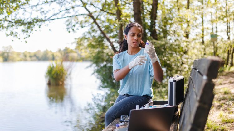 Jeune femme portant des gants réalise des recherches environnementales près d'un lac. Elle utilise du matériel, y compris un ordinateur portable et des kits de test. Des arbres et de l'eau en arrière-plan.