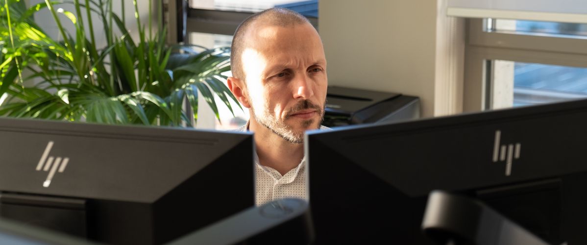The image shows a man sitting at a desk, focused on two computer screens in front of him. He has short hair and a well-groomed beard. He is wearing a white shirt with subtle patterns. In the background, there is a green plant that adds a touch of nature to the work environment. Natural light is coming through a window, illuminating the scene.