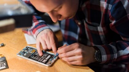An individual is working on the inside of a phone, using a tool to manipulate electronic components. A wooden table is visible, with spare parts next to it.