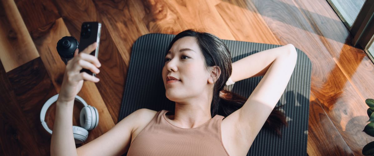 A woman lying on a yoga mat, looking at her phone with headphones beside her. Natural light illuminates the wooden room.