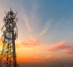 A metal telecommunications tower with multiple antennas stands against a colorful sky painted with orange, pink, and blue hues during sunset.