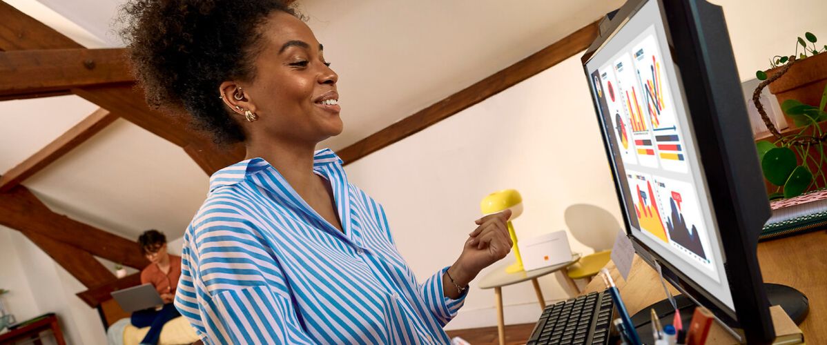 Une femme souriante est assise à un bureau dans un espace de télétravail aménagé sous les combles. Elle travaille sur un ordinateur de bureau affichant des graphiques et des tableaux de données colorés. Elle tient un stylo et prend des notes, tandis qu’une autre personne travaille en arrière-plan dans la même pièce.