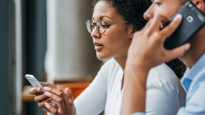 Two people using smartphones: one looking at the screen while the other is making a phone call.