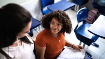 Two young women are sitting in a classroom. One of them is holding an open document on her lap while they look at each other and talk. Blue school desks and chairs are visible around them.