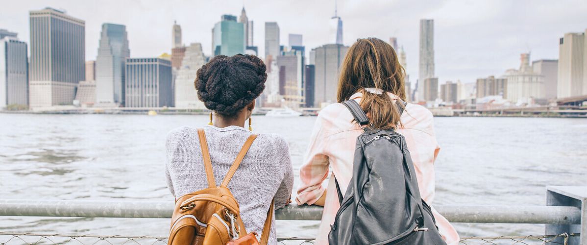 Deux femmes portant des sacs à dos regardent l’horizon depuis une promenade au bord de l’eau, face à une grande ville et ses gratte-ciel.