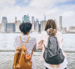 Deux femmes portant des sacs à dos regardent l’horizon depuis une promenade au bord de l’eau, face à une grande ville et ses gratte-ciel.