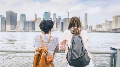 Two women wearing backpacks look at the skyline of a large city from a waterfront promenade.