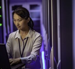 Une femme travaille sur un ordinateur portable au sein d’un data center. Elle se tient entre des rangées de serveurs informatiques éclairés par des voyants lumineux bleus et verts, dans un environnement technique et sécurisé.