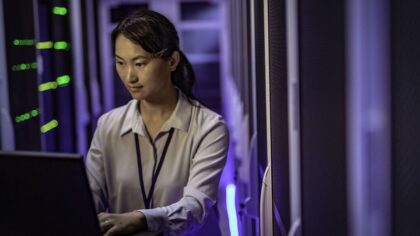 A woman is working on a laptop inside a data center. She is standing between rows of server racks illuminated by blue and green indicator lights, in a technical and secure environment.