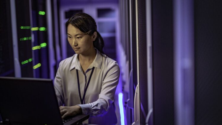 A woman is working on a laptop inside a data center. She is standing between rows of server racks illuminated by blue and green indicator lights, in a technical and secure environment.