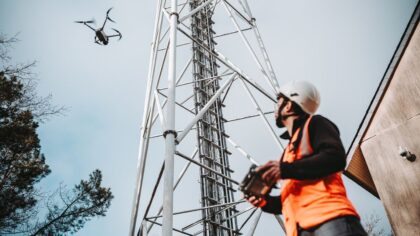 A technician wearing a helmet and safety vest operates a drone near a telecommunications tower.