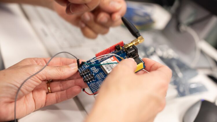 Alt text: Hands are handling an electronic circuit board fitted with components, wires, and an antenna. The person is connecting or adjusting parts of the device on a technical work surface.